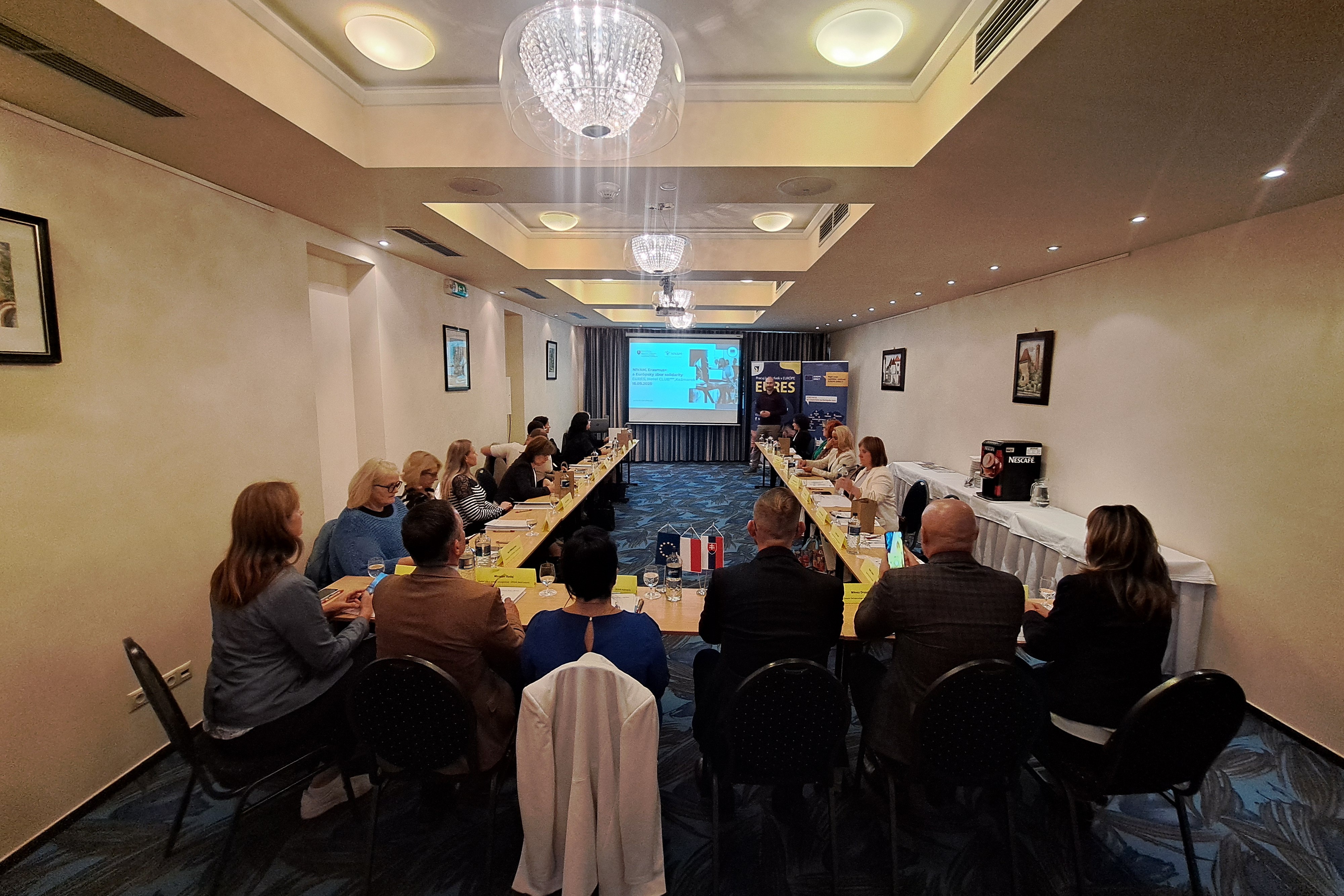 Sala konferencyjna pełna elegancko ubranych uczestników, siedzących przy stołach i wsłuchujących się w prezentację