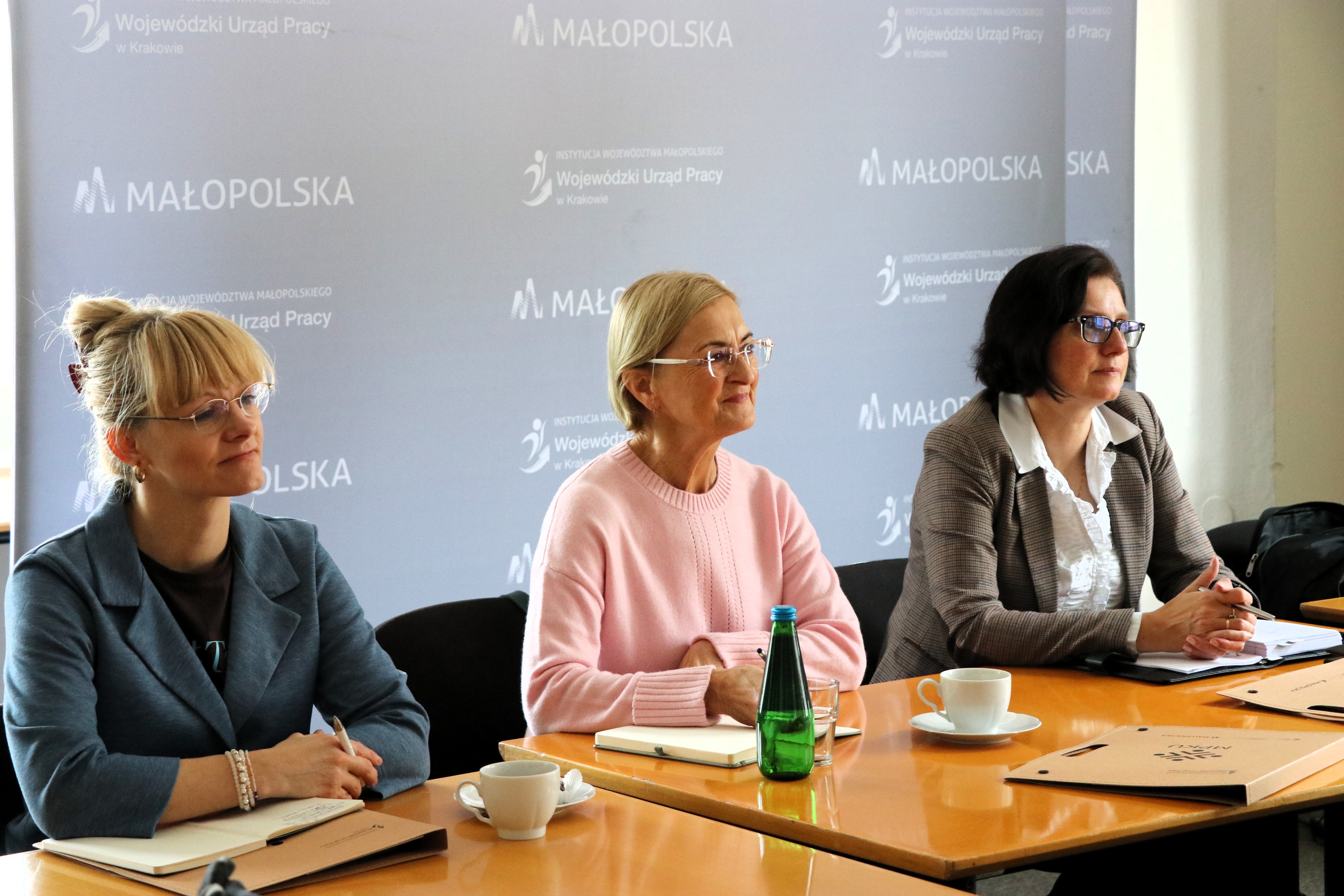Trzy kobiety siedzą przy stole. Słuchają. W tle ścianka Wojewódzkiego Urzędu Pracy w Krakowie.