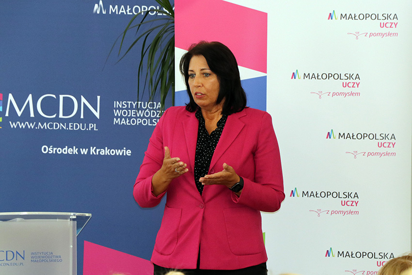 Kobieta  w różowej marynarce, gestykuluje i mówi. W tle roll up Małopolskiego Centrum Doskonalenia Nauczycieli i Małopolska Uczy.