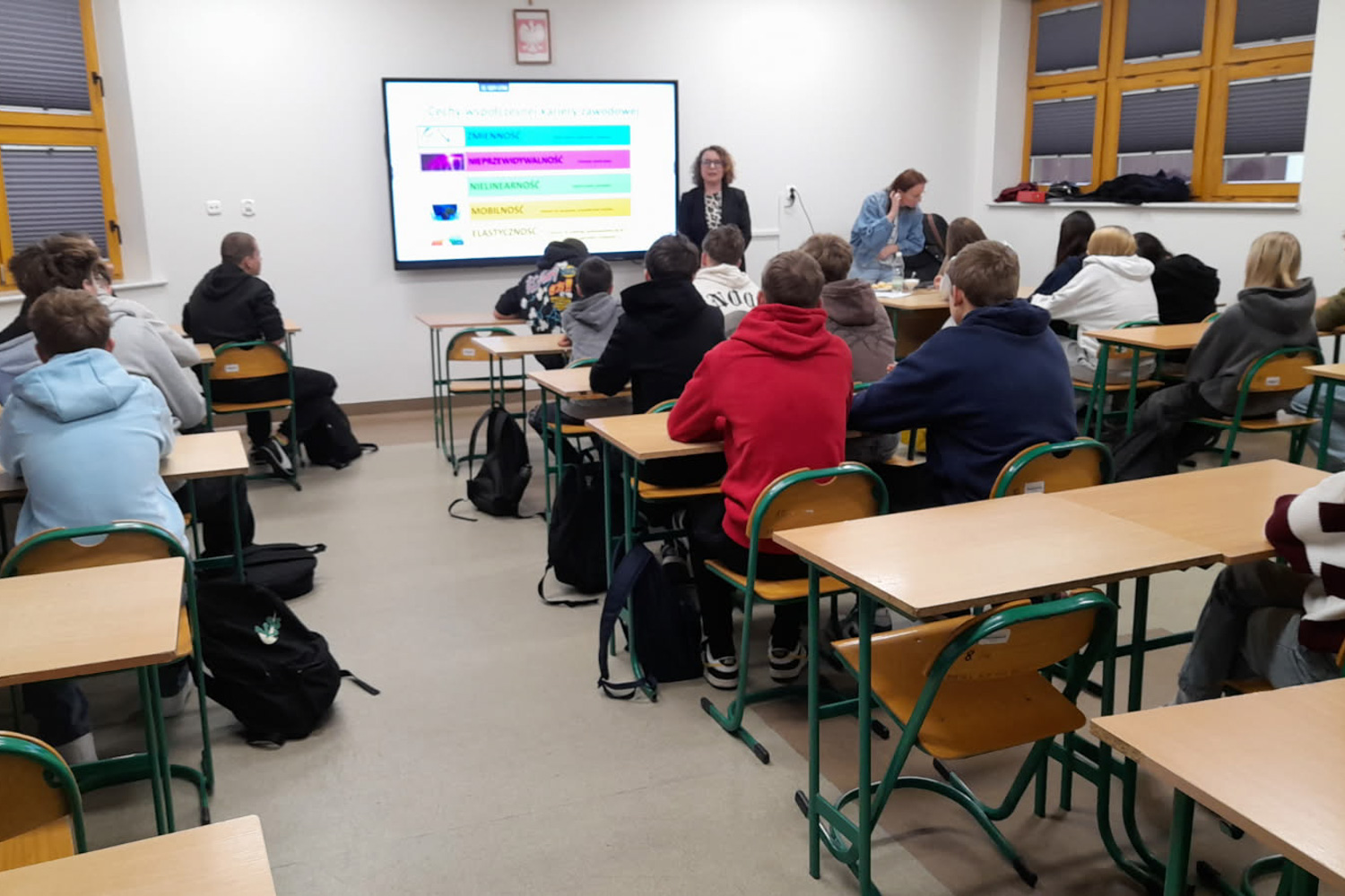 Sala lekcyjna w szkole. W pomieszczeniu siedzi kilkunastu nastolatków, zwróconych twarzami w stronę tablicy multimedialnej, na której wyświetlona jest kolorowa prezentacja.