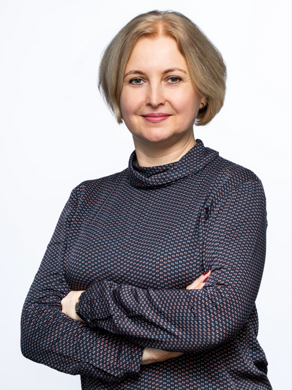 Uśmiechnięta kobieta, z krótkimi blond włosami, stoi z założonymi na siebie rękoma